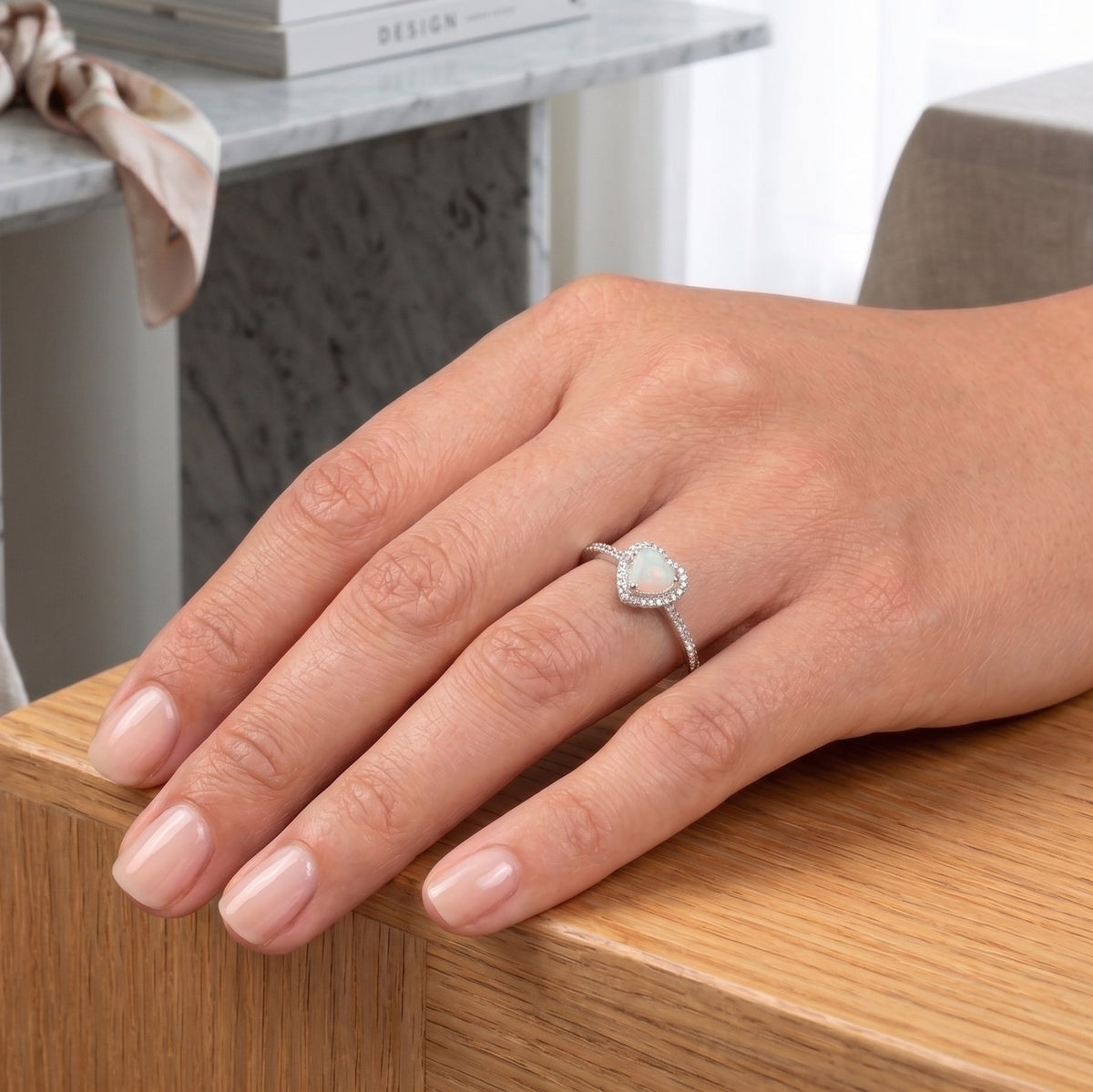 A close-up of a hand wearing a silver ashes memorial ring with a heart-shaped opal ashes stone, resting on a wooden table in a softly lit interior setting.