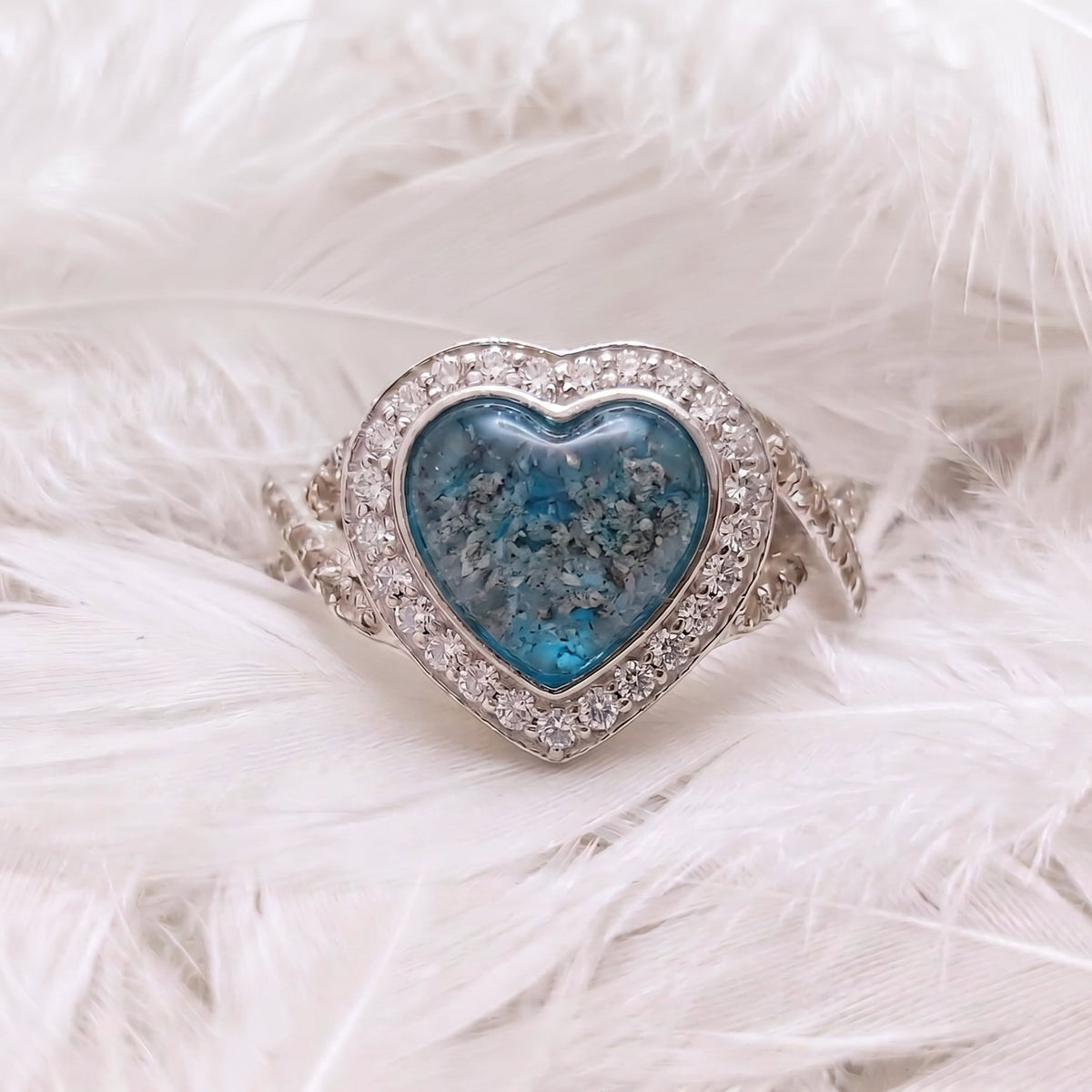 A heart-shaped silver ashes memorial ring featuring a central blue topaz ashes stone, surrounded by a halo of cubic zirconia stones, displayed on a soft white feather background.