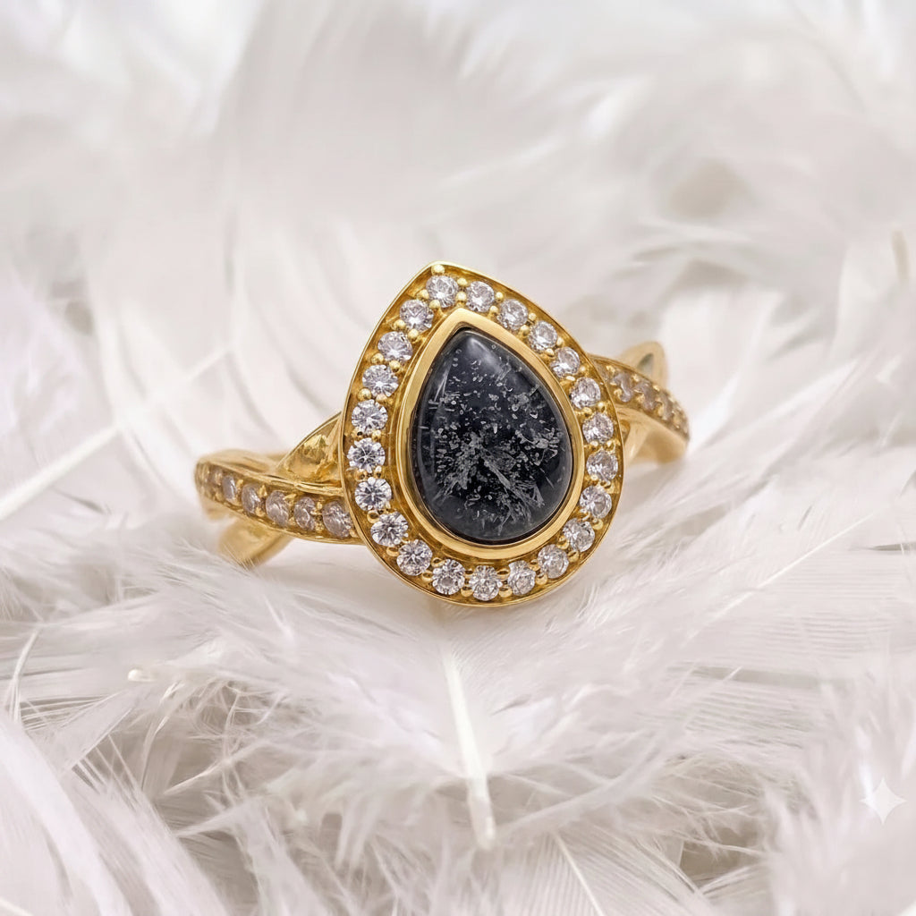 18ct gold ashes memorial ring featuring a teardrop-shaped black ashes stone surrounded by a halo of real diamonds, displayed on a bed of white feathers.
