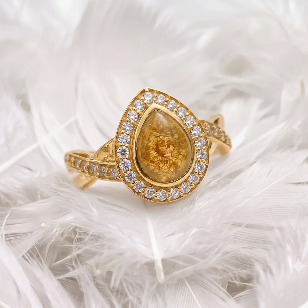 14ct or 18ct gold ashes ring featuring a teardrop-shaped ashes gemstone, surrounded by a halo of real diamonds, resting on white feathers.