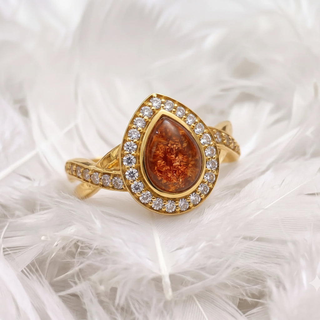 18ct gold ashes memorial teardrop-shaped ring with ambber-orange ashes stone and a halo of real diamonds, resting on white feathers.