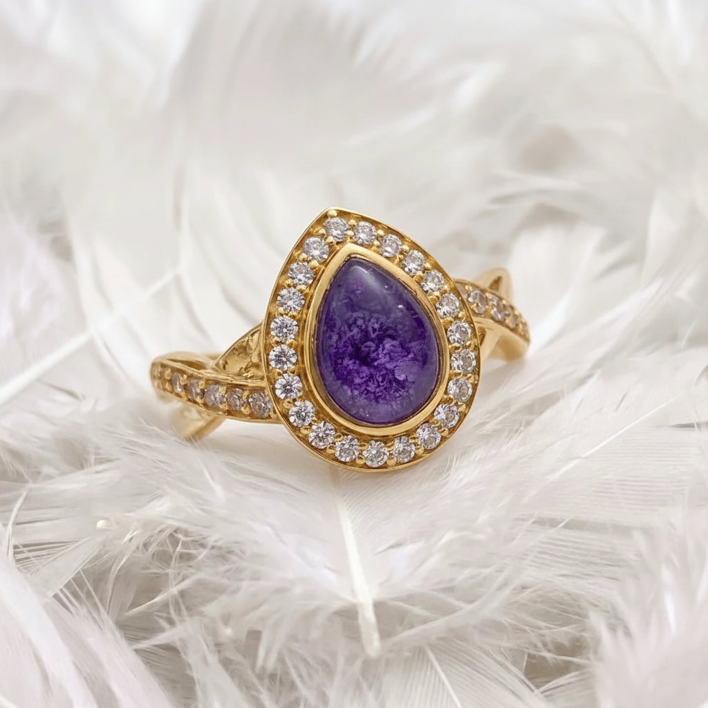 14ct or 18ct gold ashes memorial ring teardrop-shaped ring featuring a  purple ashes stone, surrounded by a halo of real diamonds, resting on white feathers.