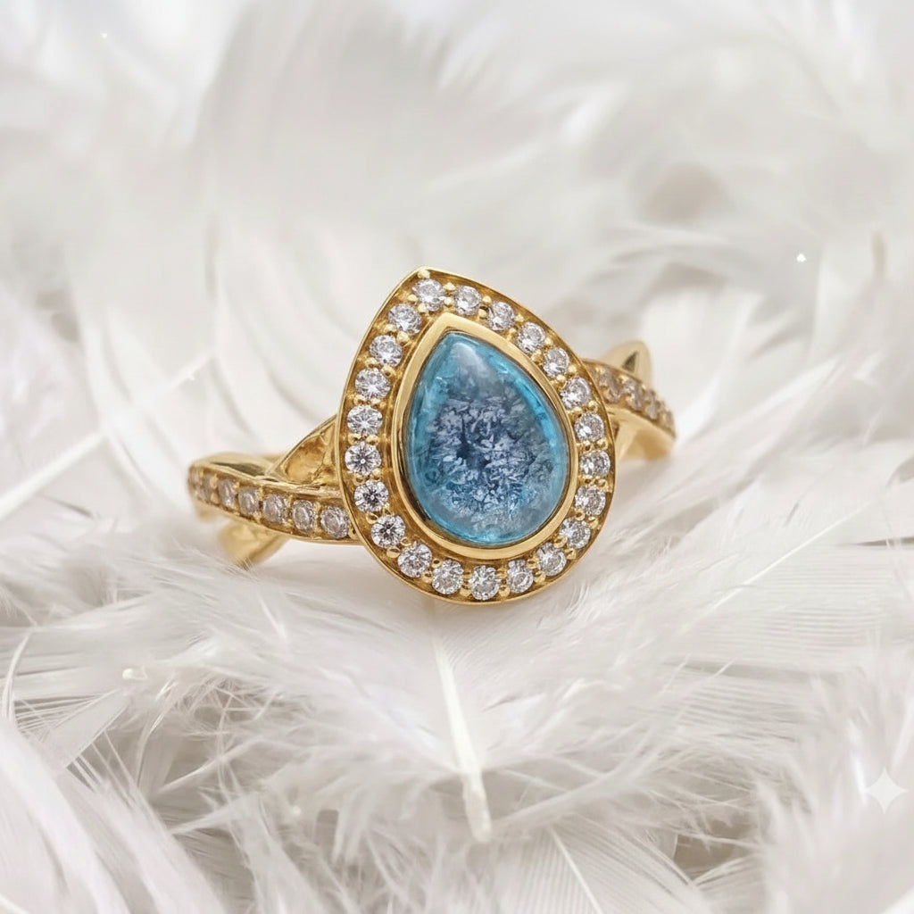 14ct or 18ct gold teardrop-shaped ashes ring surounded by a halo of real diamonds, resting on white feathers.