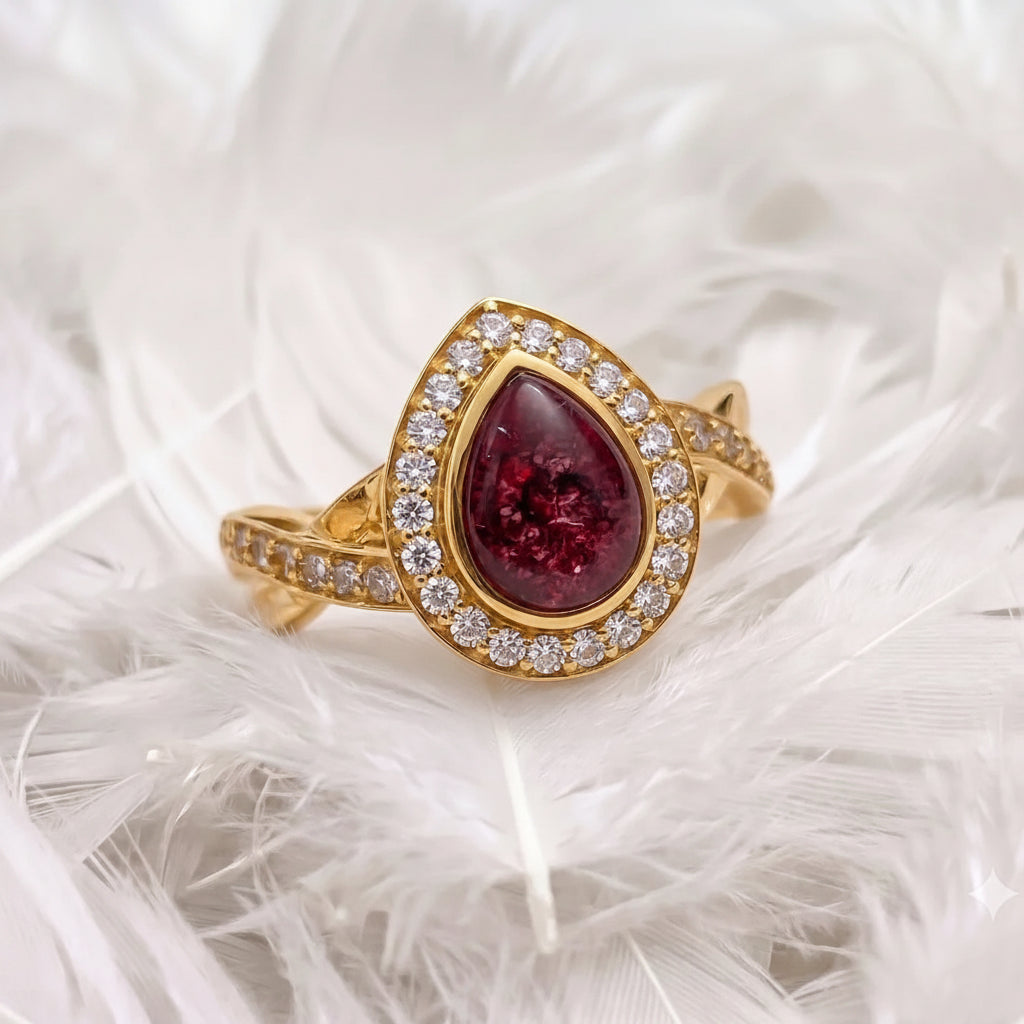14ct or 18ct gold ashes ring featuring a teardrop-shaped deep red ashes stone surrounded by a halo of real diamonds, displayed on a bed of white feathers.