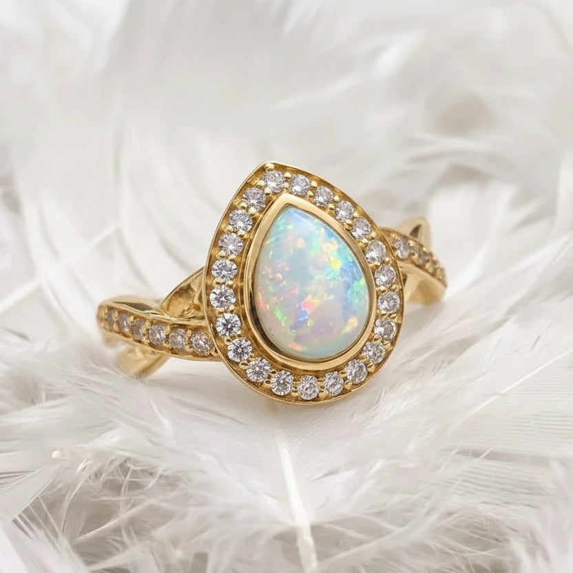 14ct gold ashes memorial ring featuring a pear shaped  iridescent opal ashes stone surrounded by a halo of real diamonds, resting on white feathers.
