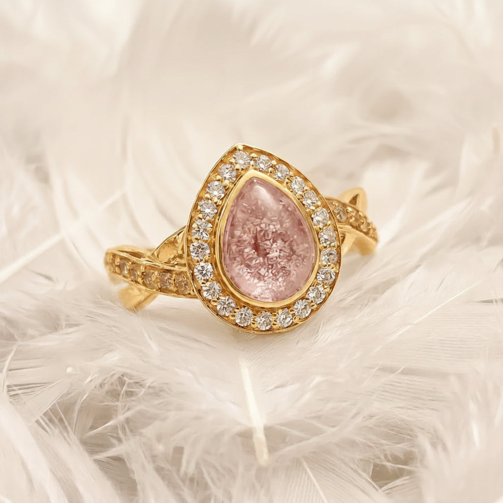 14ct or 18ct gold ashes memorial ring featuring a teardrop-shaped pink topaz ashes stone surrounded by a halo of real diamonds, displayed on a bed of white feathers.