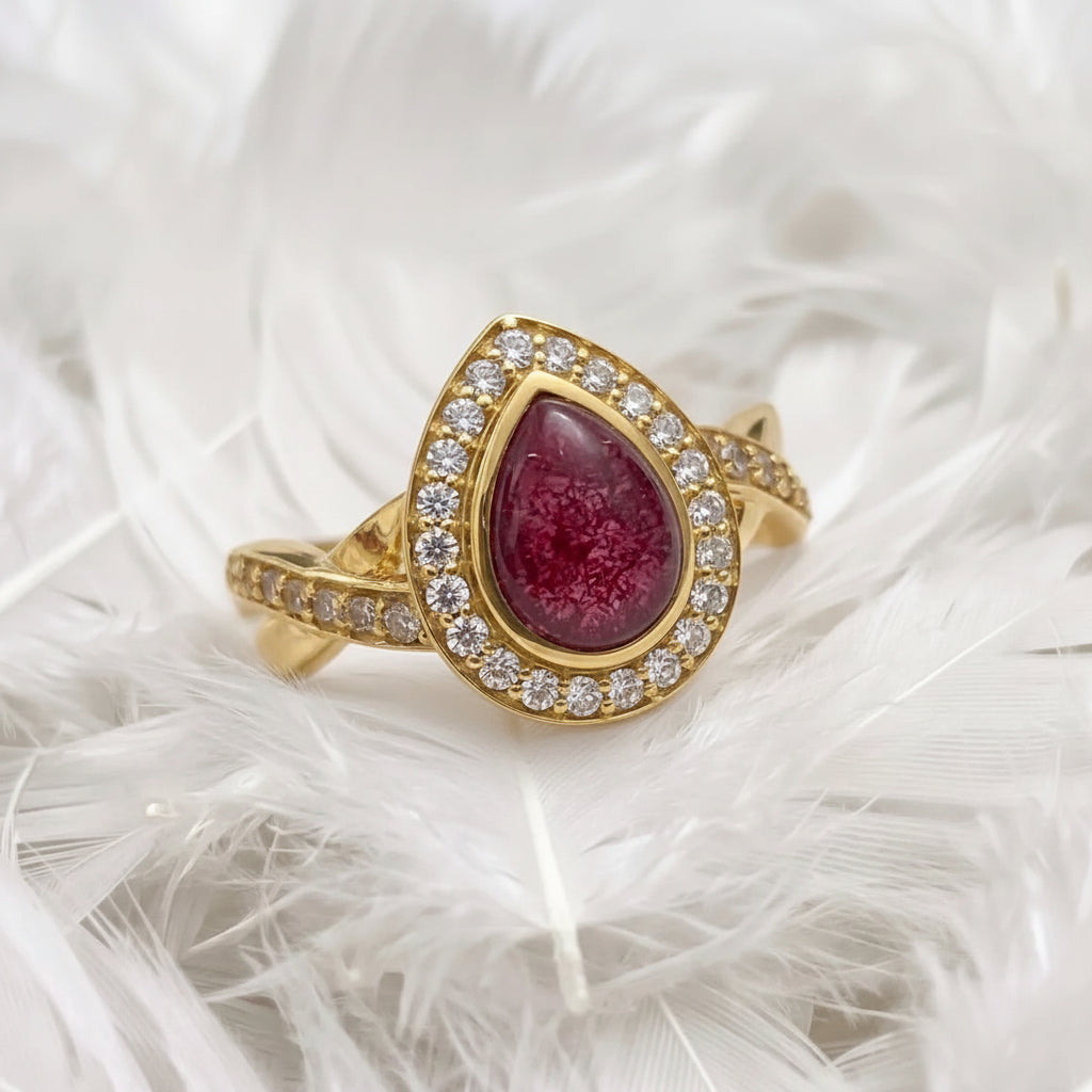 14ct gold teardrop-shaped ashes memorial ring featuring a garnet ashes stone surrounded by a halo of real diamonds, displayed on a bed of white feathers.