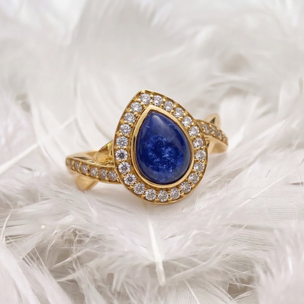 18ct gold ashes memorial ring with a teardrop-shape blue sapphire ashes stone surrounded by a halo of real diamonds, displayed on a bed of white feathers.