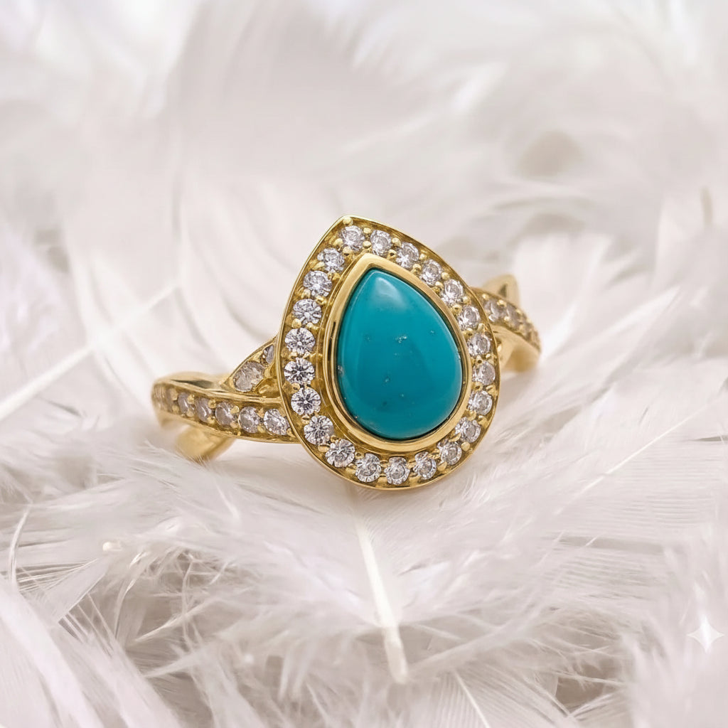 14ct gold ashes memorial ring featuring a teardrop-shaped turquoise ashes stone surrounded by a halo of real diamonds displayed on a bed of white feathers.