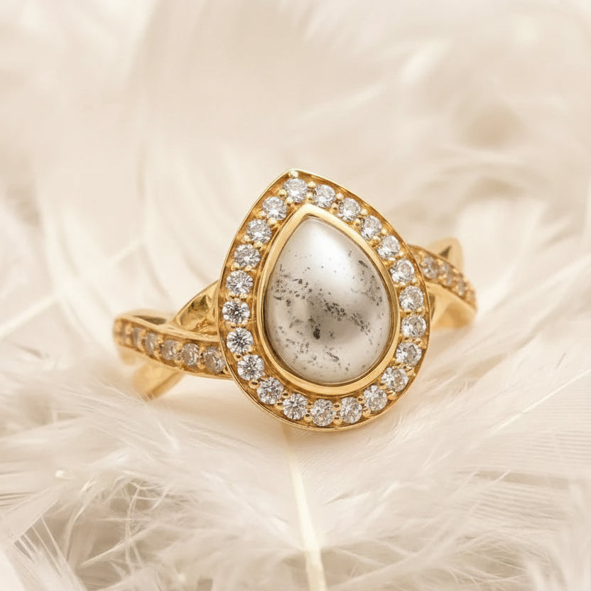18ct gold ashes memorial ring with teardrop-shaped ashes stone in a pearl colour and a halo of real diamonds, displayed on white feathers.
