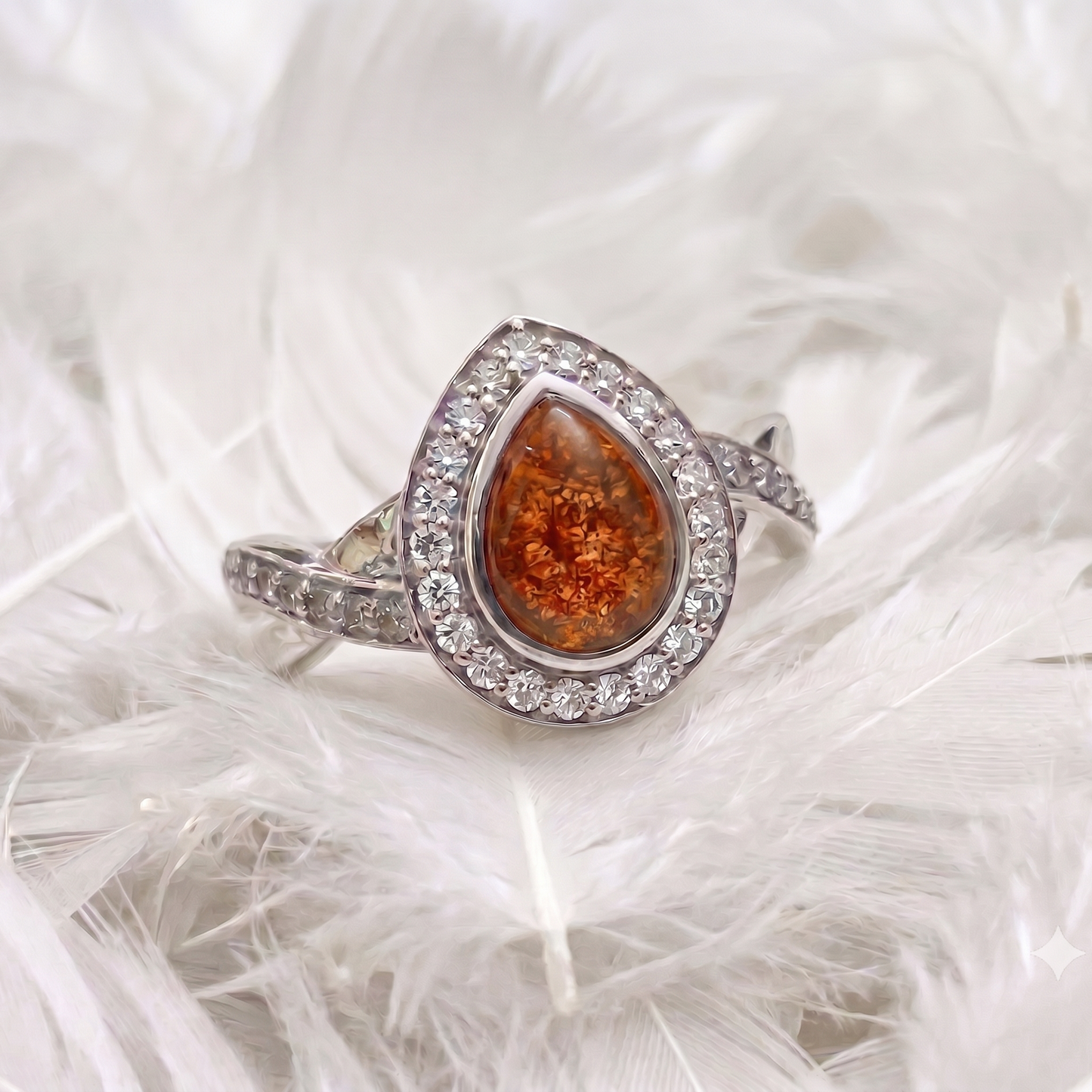 A silver teardrop-shaped ring with a sparkling amber-orange center stone and a halo of small clear stones, resting on white feathers.