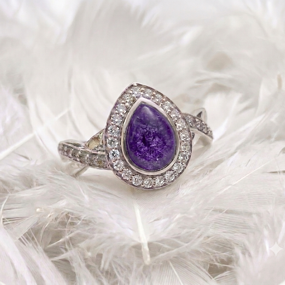 A teardrop-shaped purple ashes  gemstone ring with a silver-toned halo and pavé band, resting on a bed of white feathers.