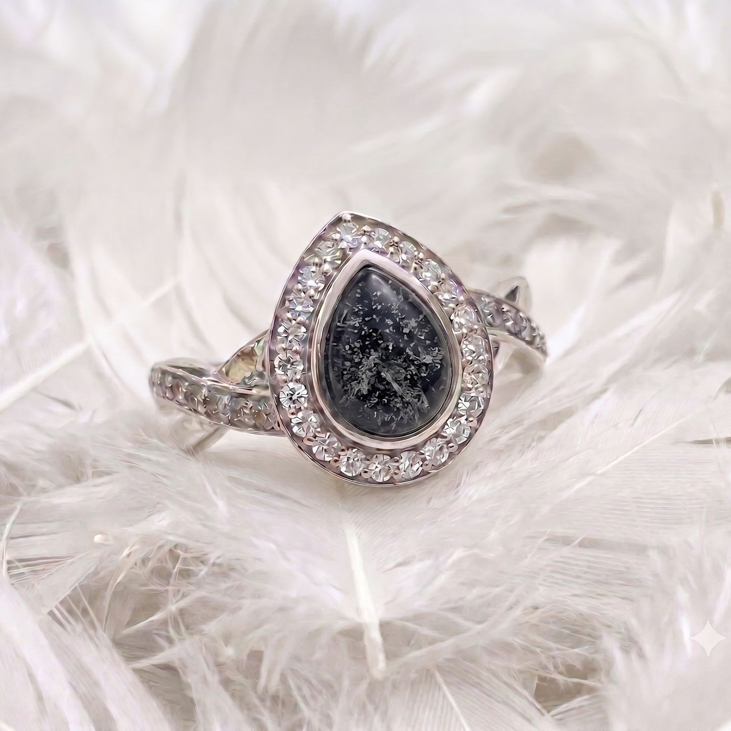 A silver ring featuring a teardrop-shaped black stone with sparkling inclusions, surrounded by a halo of small clear stones and a pavé band, displayed on white feathers.