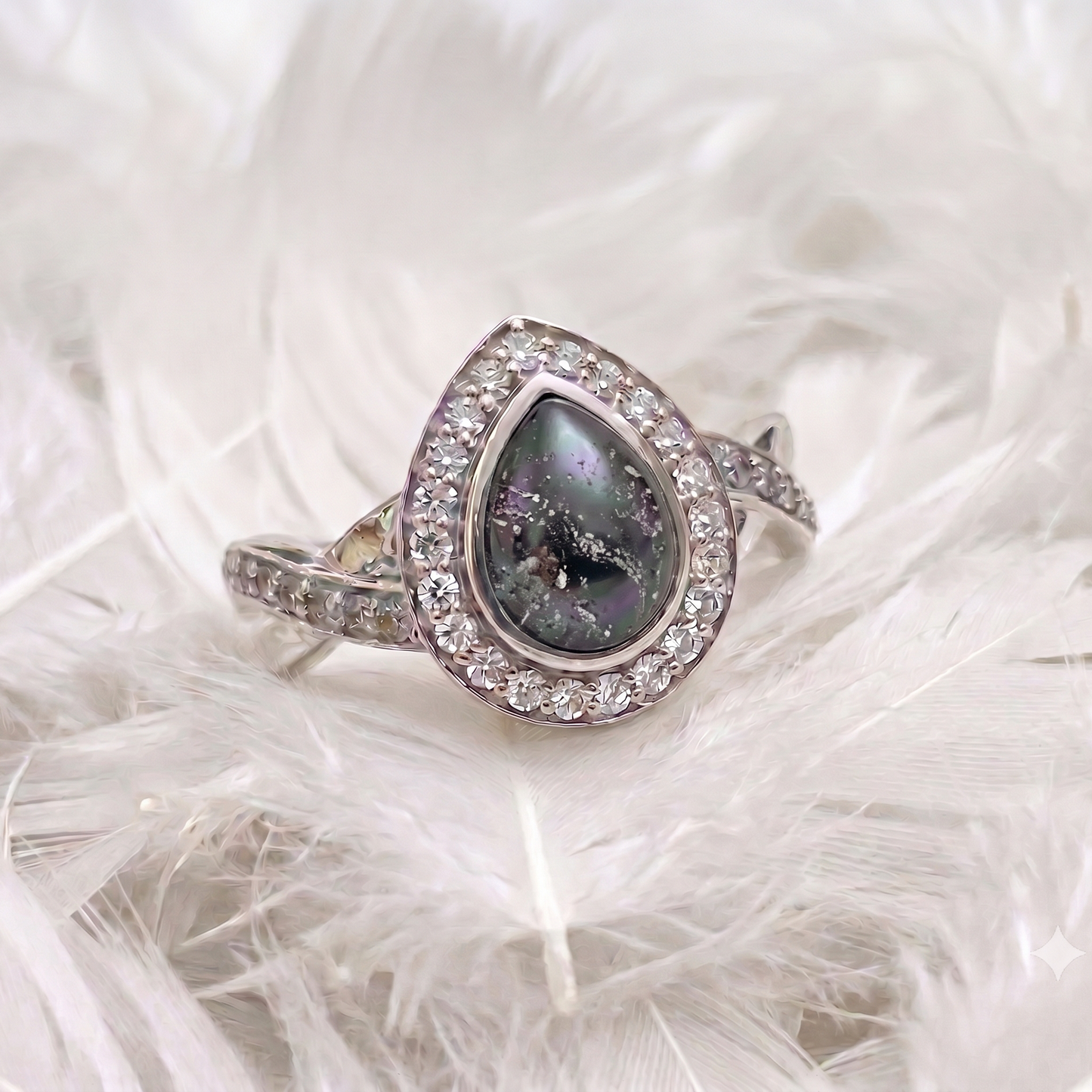 A silver-toned halo ring with a teardrop-shaped dark gray and black iridescent center stone, accented with small clear stones, resting on white feathers.