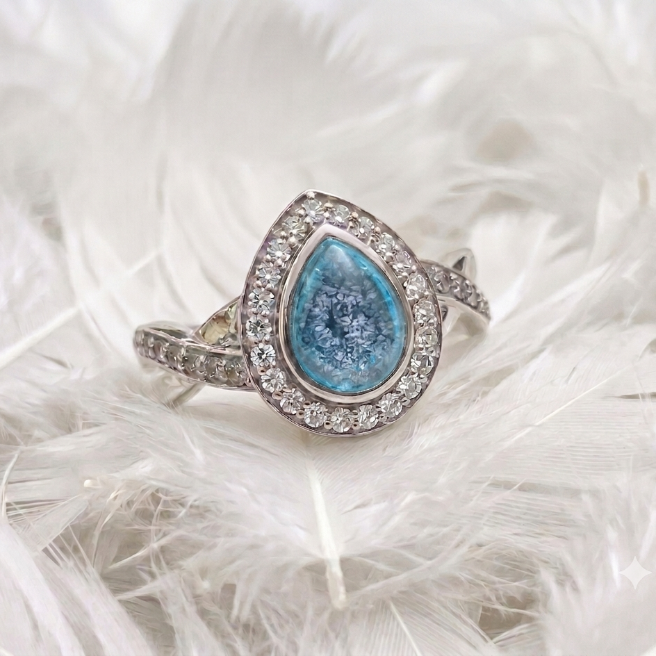 A silver ashes memorial teardrop halo ring with a textured light blue centre stone and clear pavé accents, resting on a bed of white feathers.
