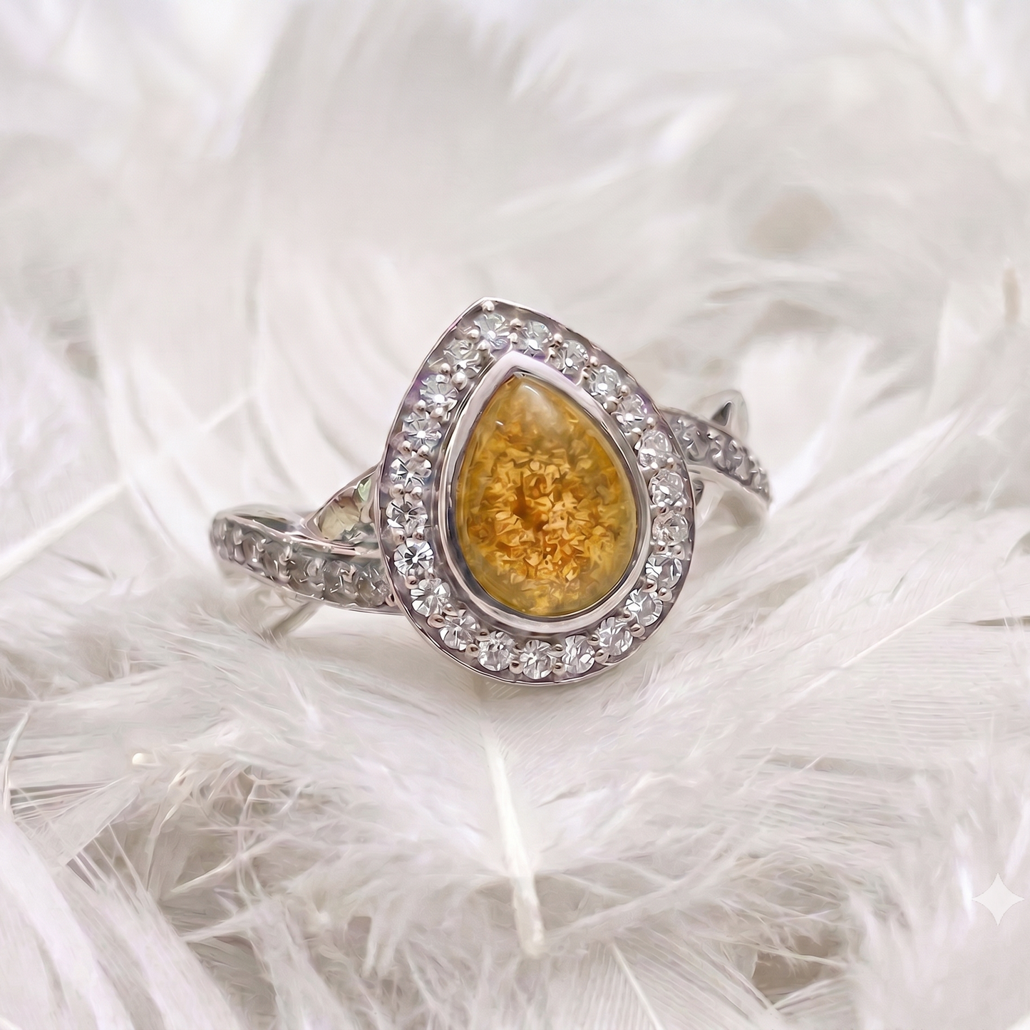 A silver ring with a teardrop-shaped yellow gold center stone, surrounded by a halo of small clear stones and a smooth outer bezel, displayed on a bed of white feathers.