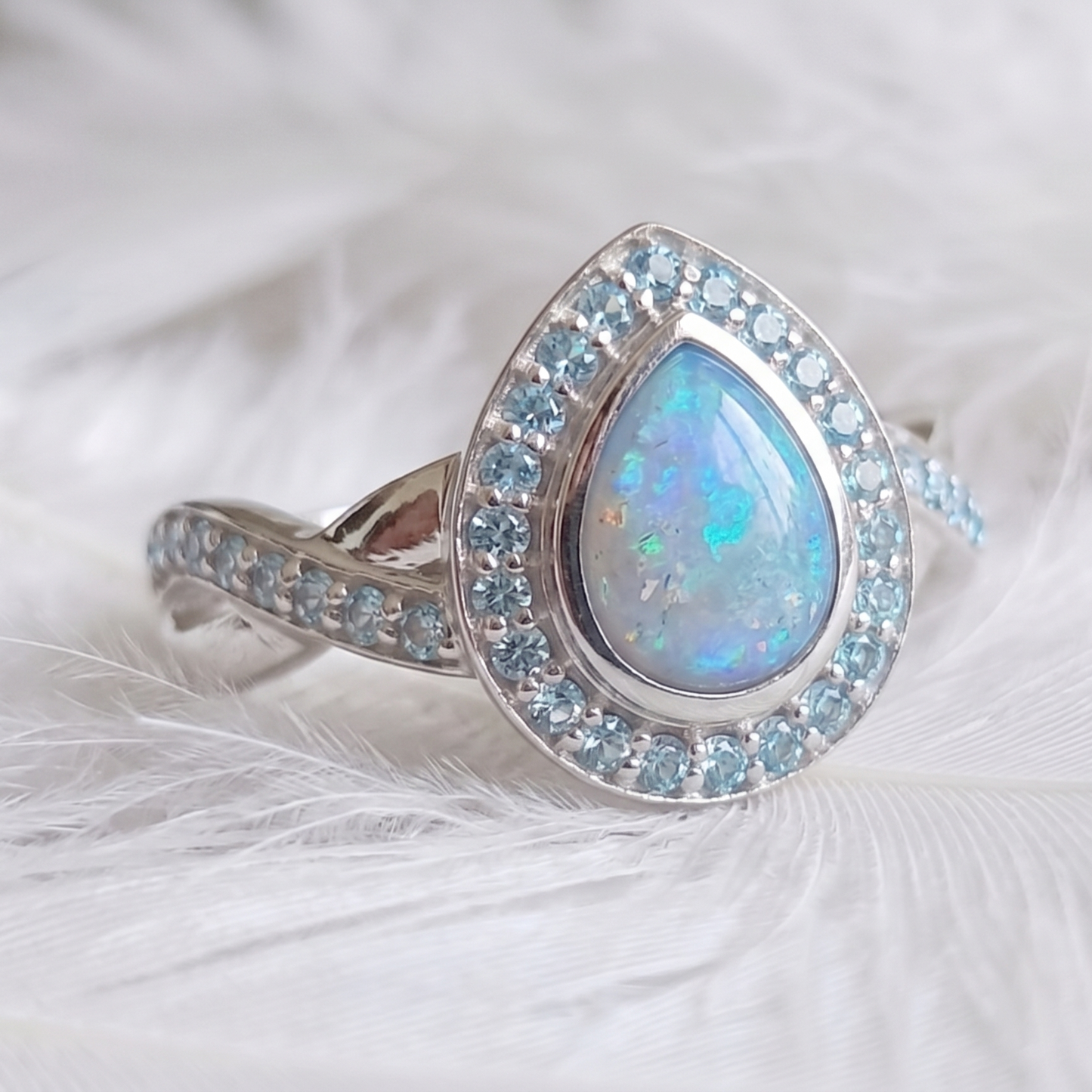 A teardrop-shaped memorial ring featuring a central iridescent blue opal surrounded by a halo of small light blue stones, set on a silver crossover twist band with pavé light blue stones, resting on white feathers.
