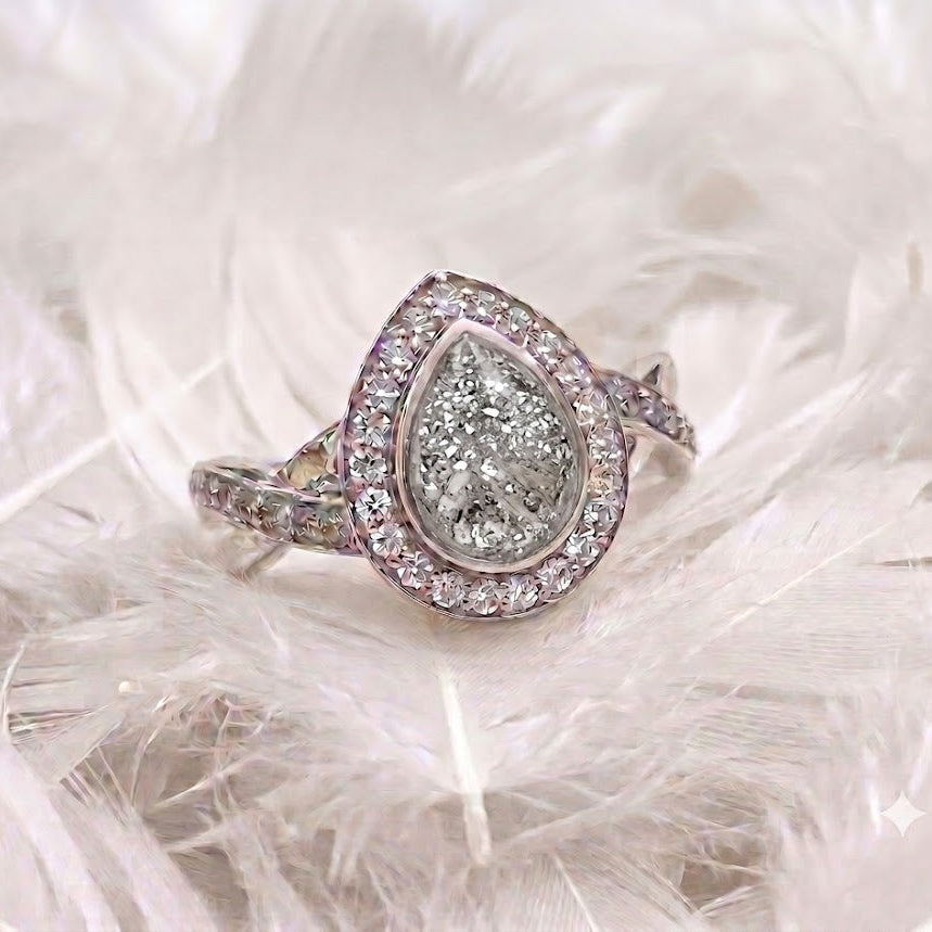 A silver teardrop-shaped ring with a halo of small clear stones and a central textured clear gemstone, displayed on a bed of white feathers.