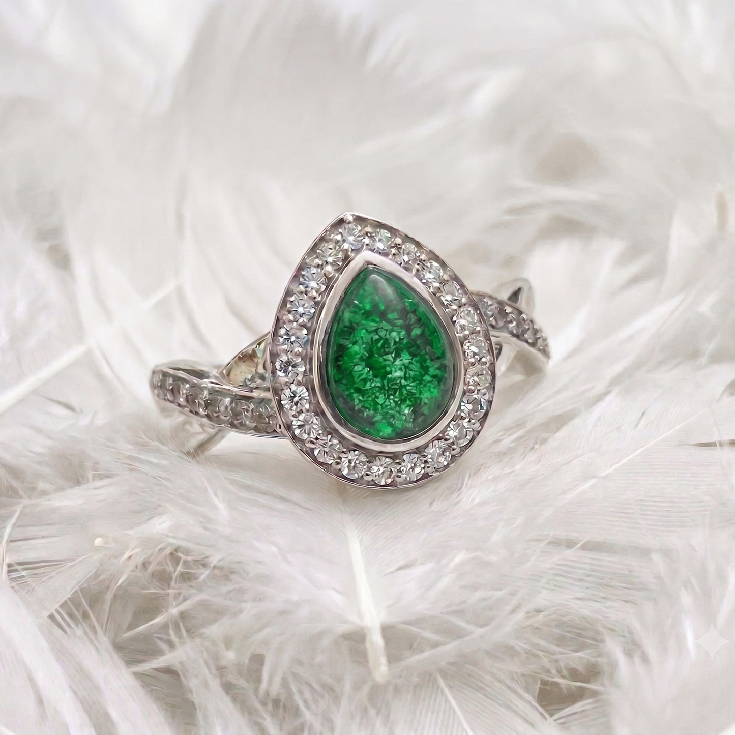 A silver ring featuring a teardrop-shaped green emerald gemstone surrounded by a halo of small clear stones, displayed on a bed of white feathers.