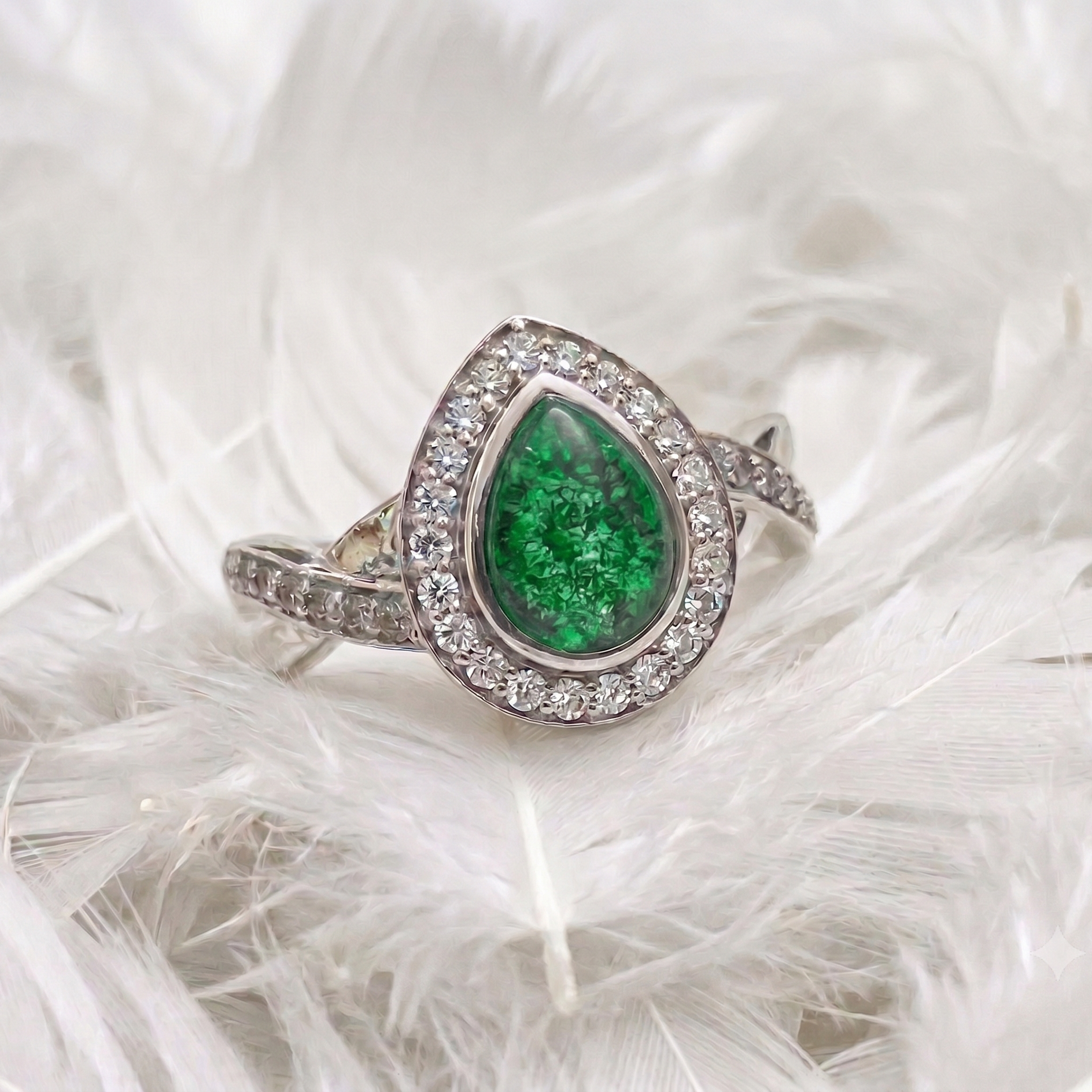 A silver ring featuring a teardrop-shaped green emerald gemstone surrounded by a halo of small clear stones, displayed on a bed of white feathers.