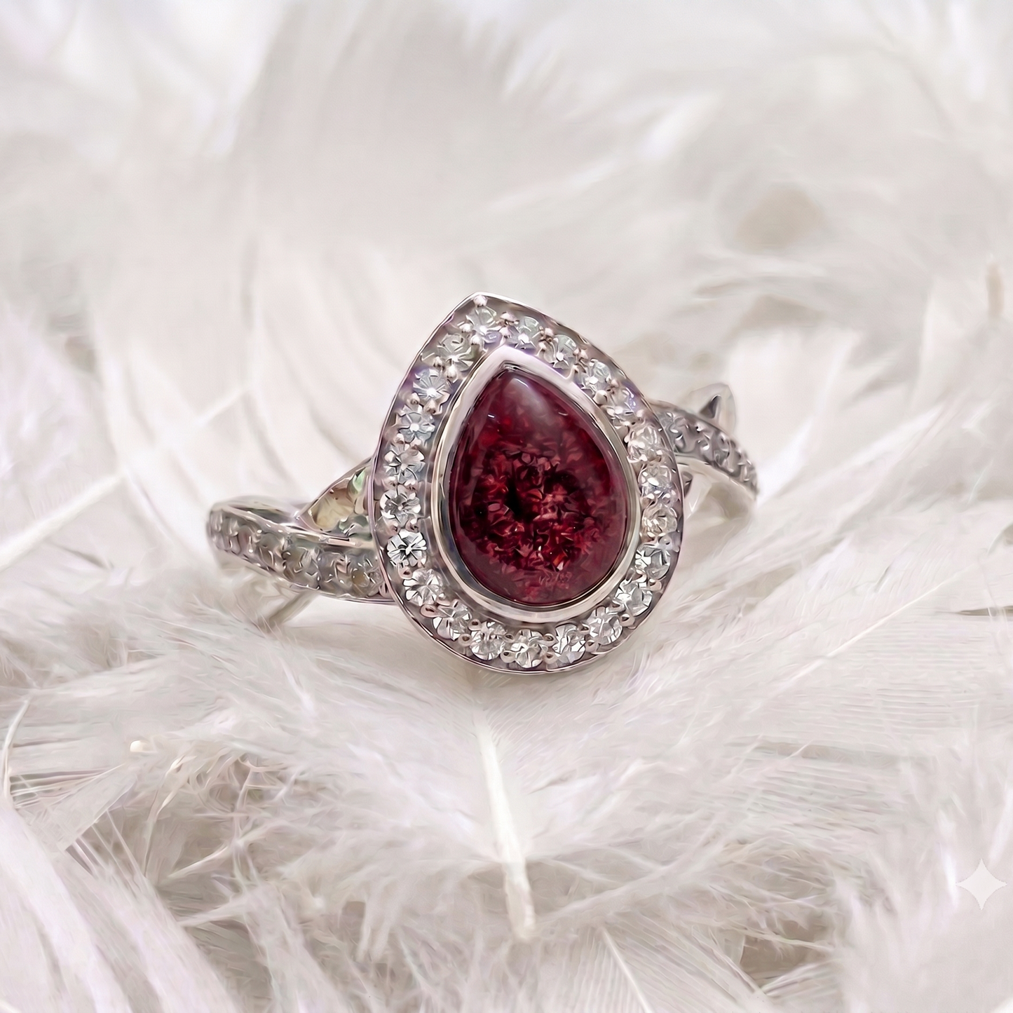 A silver ashes memorial ring featuring a teardrop-shaped deep red gemstone surrounded by a halo of small clear stones, displayed on a bed of white feathers.