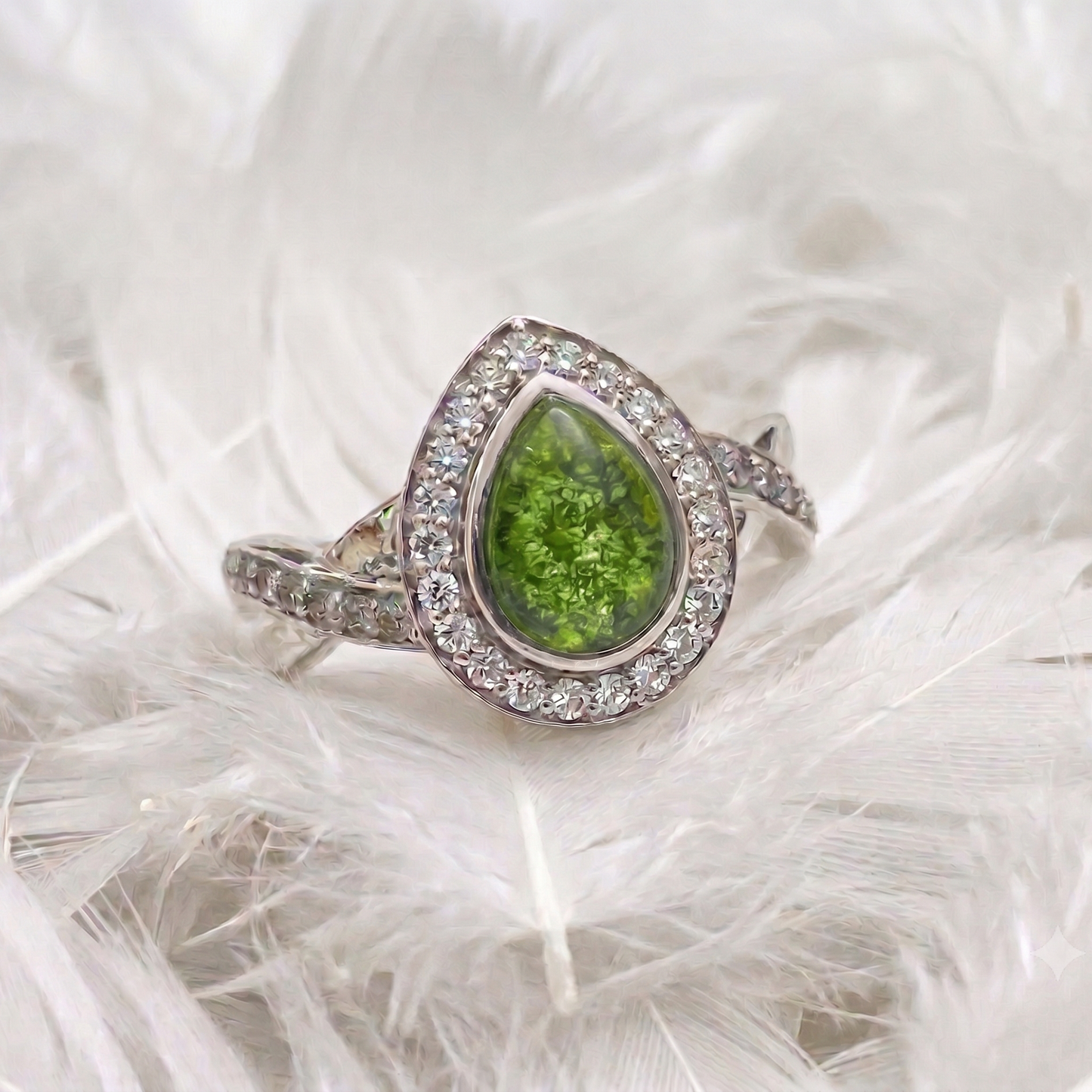 A silver ring featuring a teardrop-shaped green gemstone surrounded by a halo of small clear stones, displayed on a bed of white feathers.