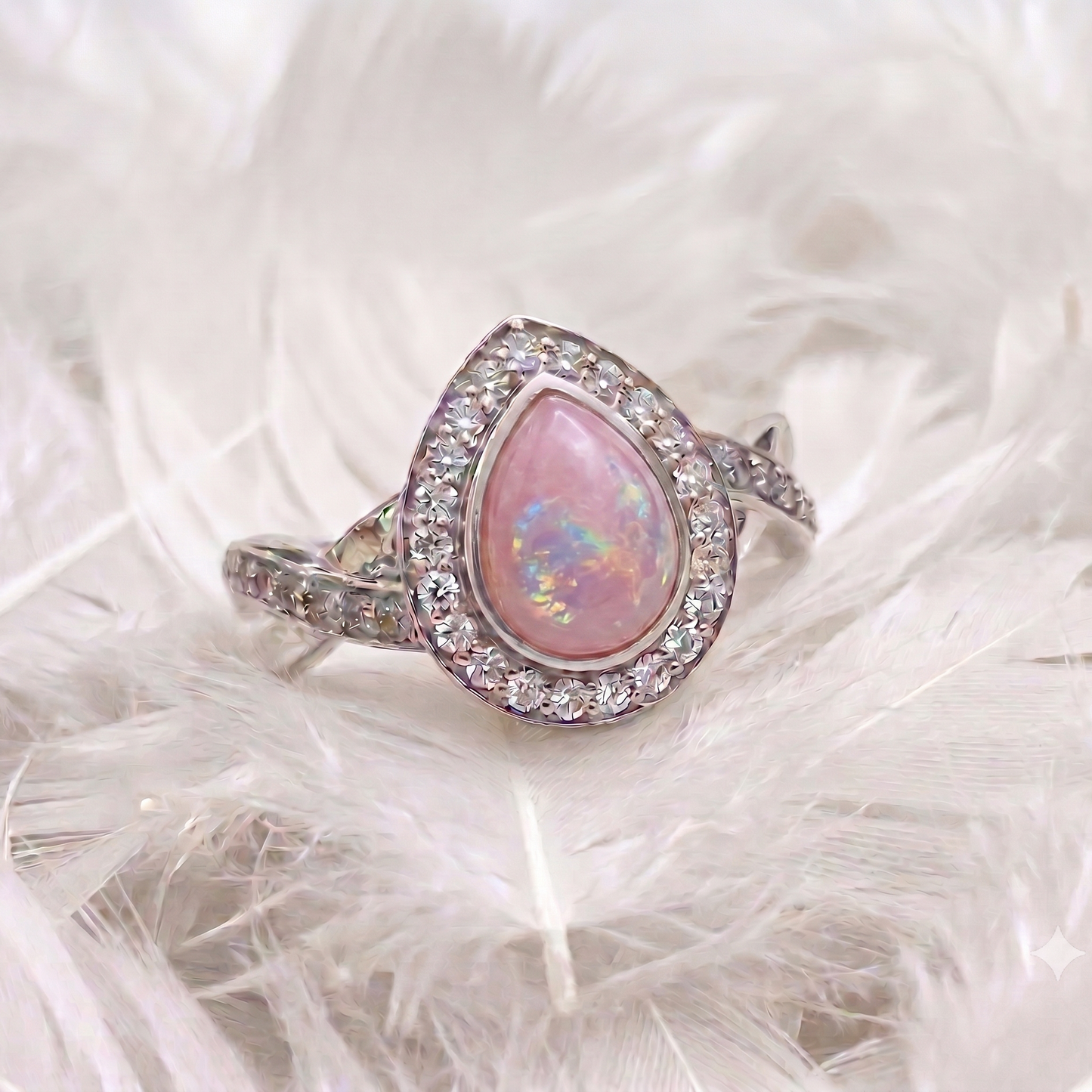 A silver ring featuring a teardrop-shaped pink opal center stone surrounded by a halo of small clear stones, displayed on a bed of white feathers.