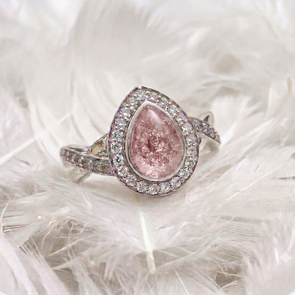 A teardrop-shaped ashes memorial pink gemstone ring with a halo of small clear stones and a silver-toned band, resting on white feathers.