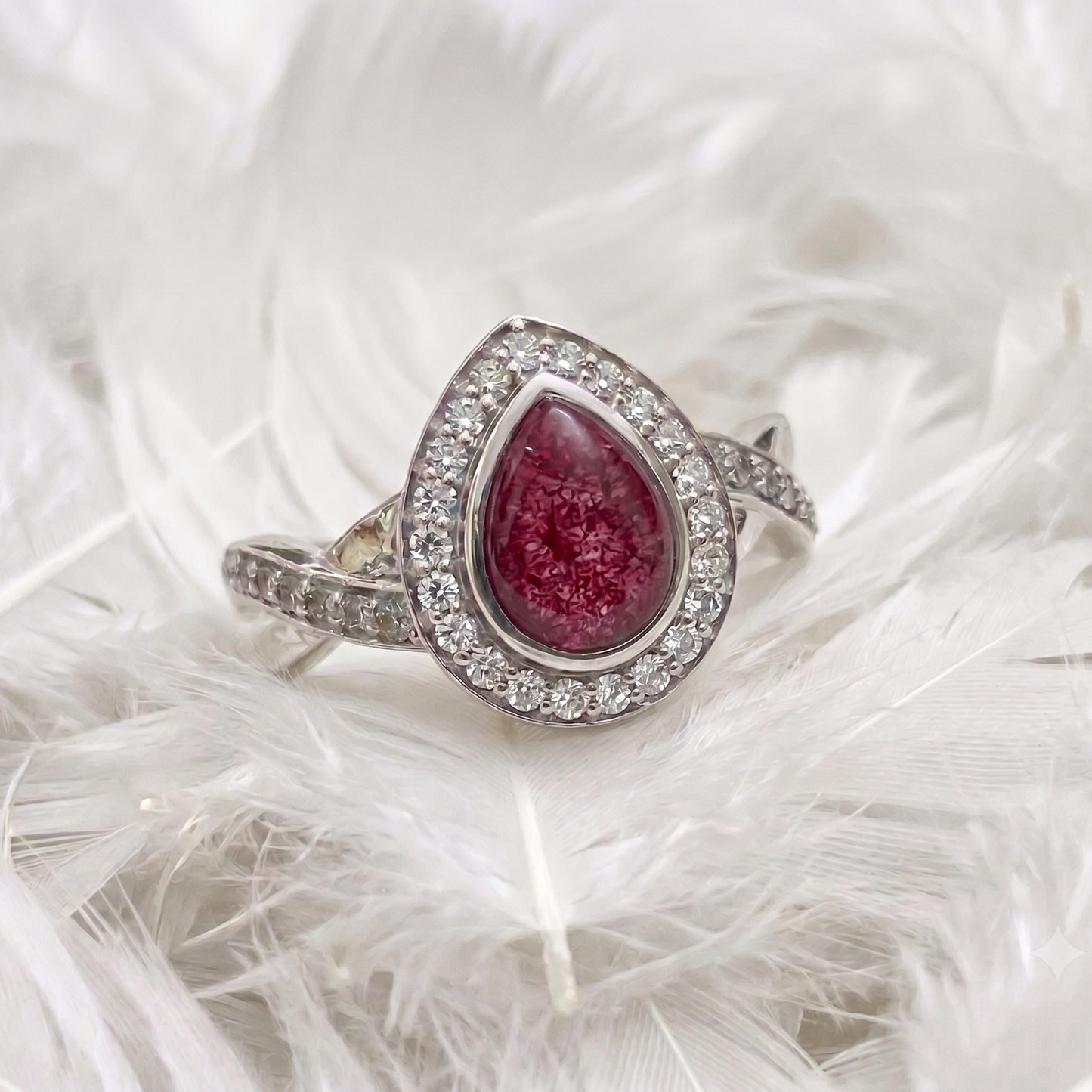 A silver ashes memorial  ring featuring a teardrop-shaped pinkish-red centre stone surrounded by a halo of small clear stones, displayed on a bed of white feathers.