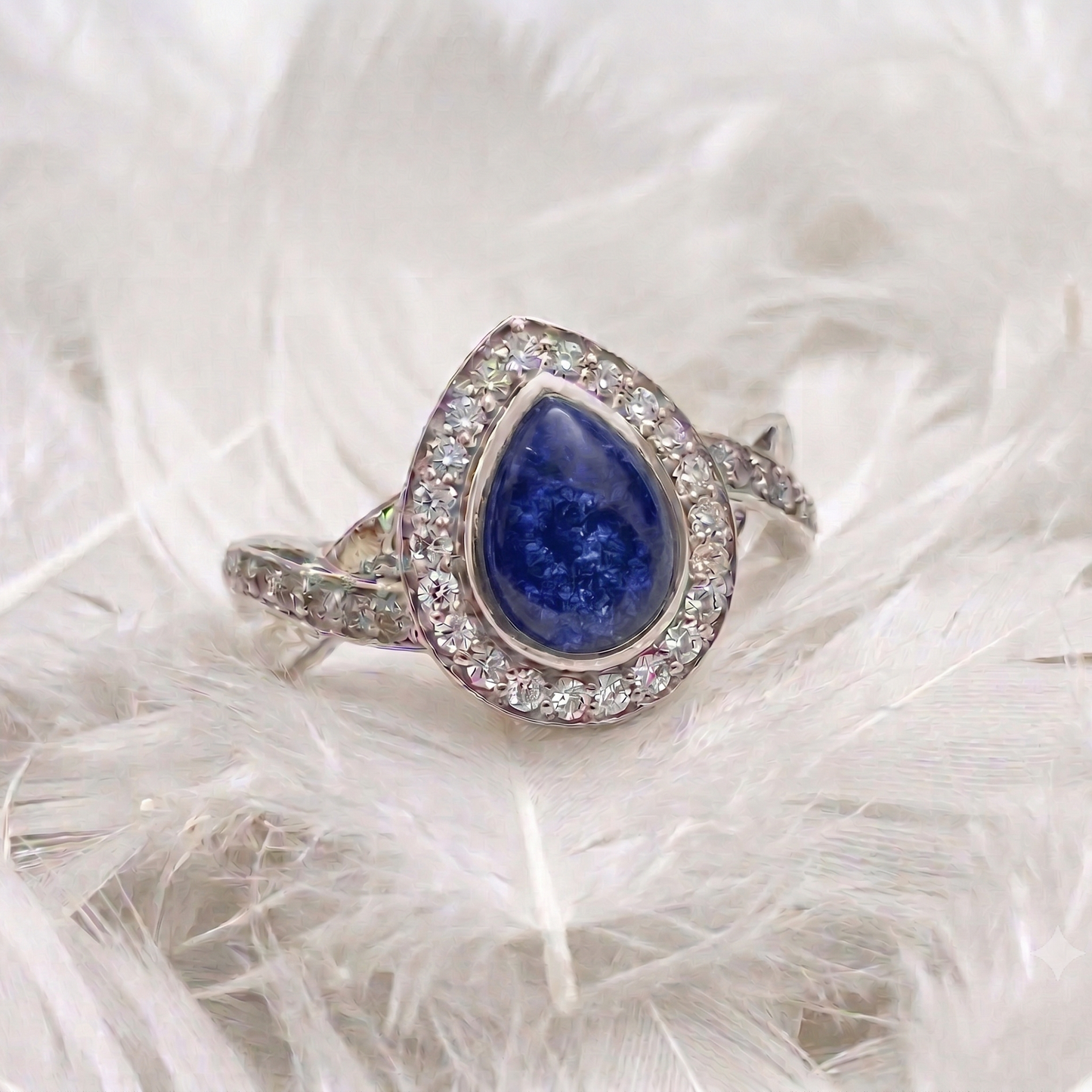 A silver ashes memorial ring featuring a teardrop-shaped blue sapphire gemstone surrounded by a halo of small clear stones, resting on a bed of white feathers