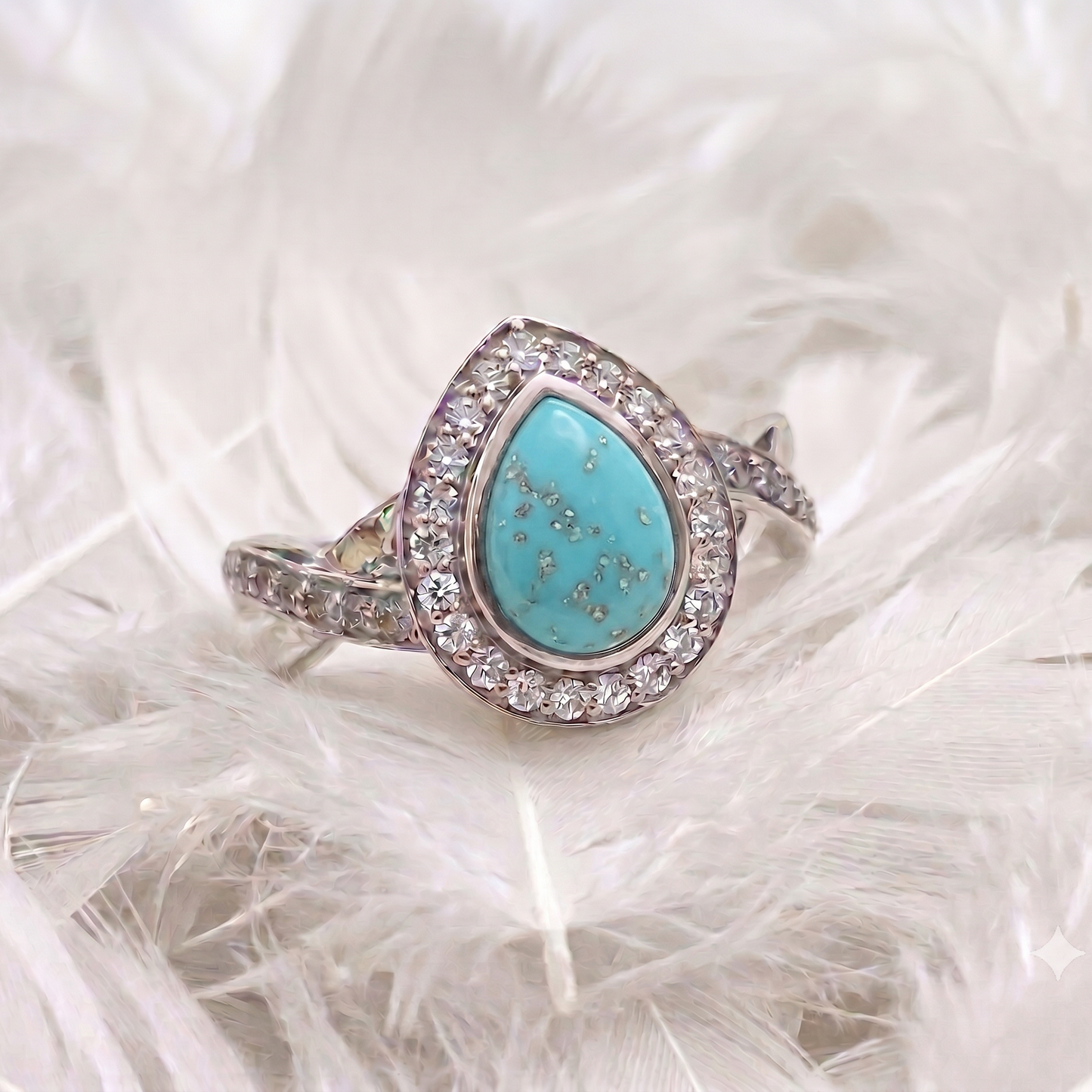 A silver ashes memorial ring featuring a teardrop-shaped turquoise stone surrounded by a halo of small clear stones, displayed on a bed of white feathers.