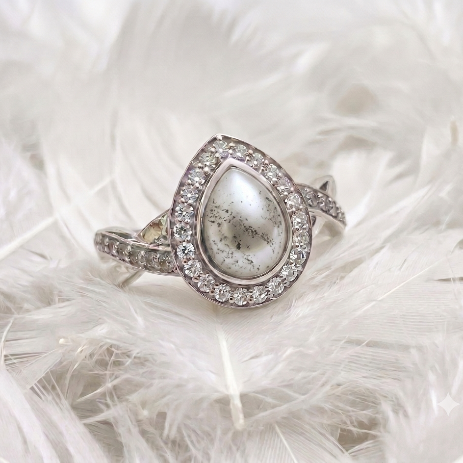 A silver-toned ring featuring a teardrop-shaped white pearl with dark inclusions, surrounded by a halo of small clear stones, resting on a bed of white feathers.
