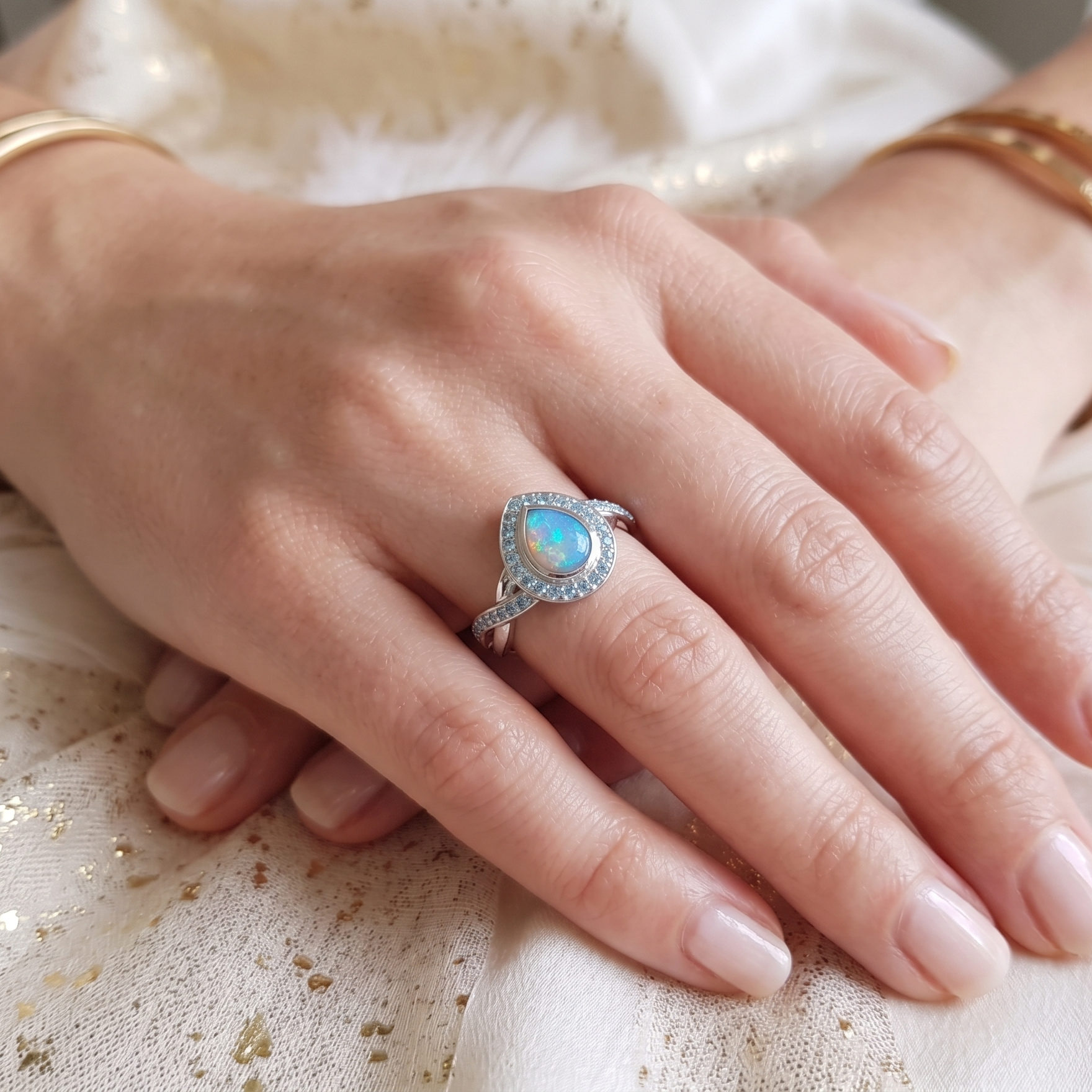 A close-up of a hand wearing a silver ring with a teardrop-shaped light blue opal center stone, surrounded by small clear stones. The hand is also adorned with gold bangles, resting on a white textured fabric with subtle gold glitter.