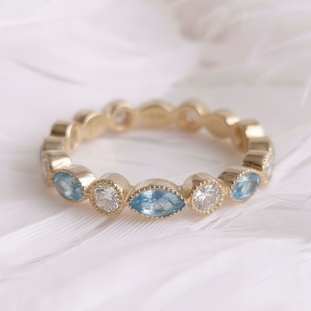 A gold eternity ring with oval-cut light blue gemstones and small clear accent stones, resting on a bed of soft white feathers.