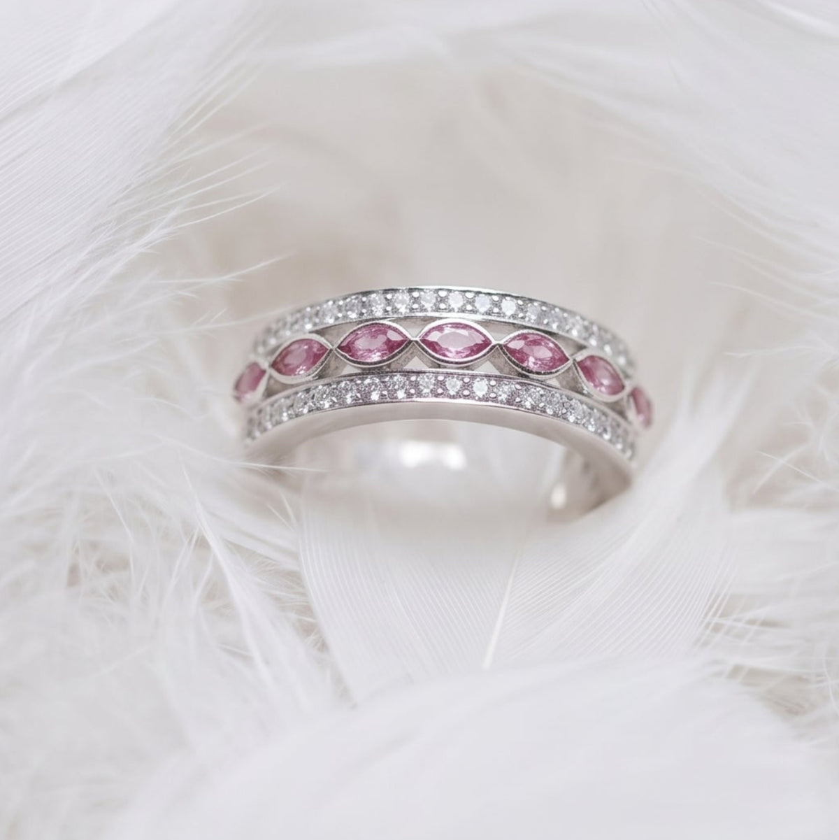 A silver wide band memorial ring with two rows of cubic zirconias and marquise ashes stones in pink, resting on a bed of soft white feathers.