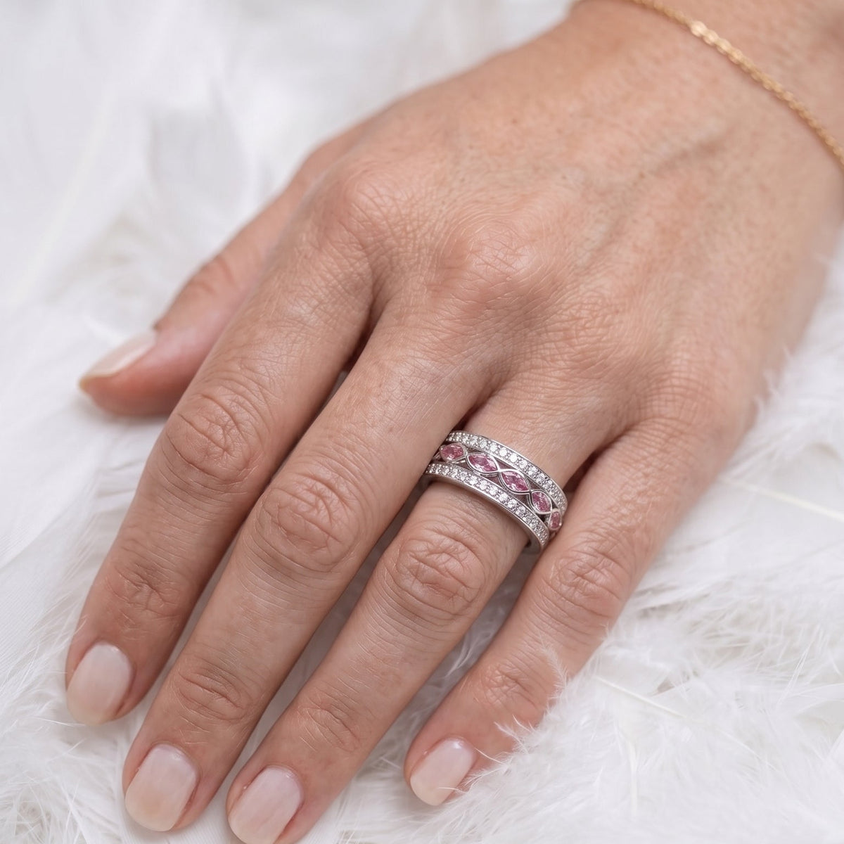 a memorial ashes ring featuring two rows of cubic zirconia on a  wide band, and marquis shape ashes stones in pink, on a background of white feathers
